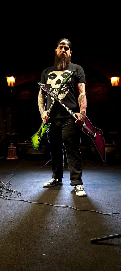 Nate Laff on a lamp-lit stage holding a green and a purple Schecter Explorer-style guitar